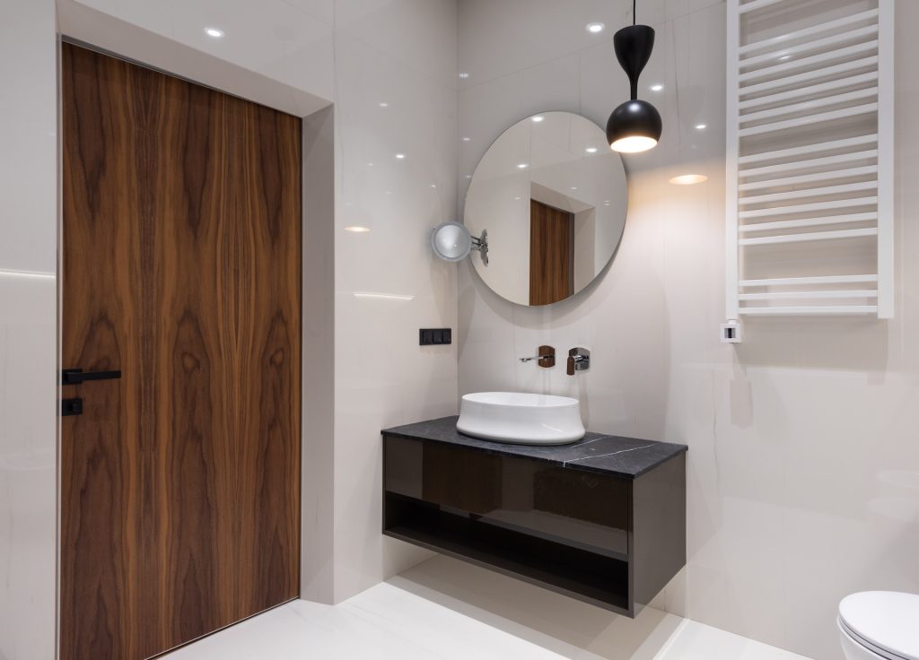 Interior of modern bright bathroom with sink on counter under round mirror near toilet and lamp hanging from ceiling near door
